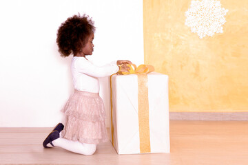 African Little girl plays with a bow color of the year 2024 peach fuzz on a large decorated gift...