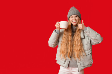 Beautiful young woman with big cup of tea showing thumb-up gesture on red background