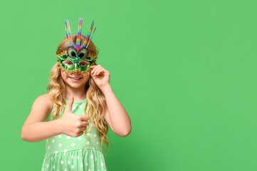 Funny little girl wearing carnival mask and showing thumb-up on green background