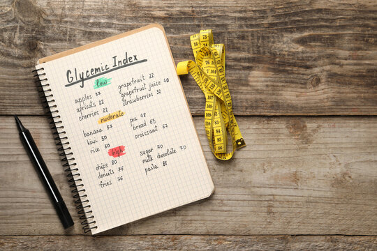 List with products of low, moderate and high glycemic index in notebook, marker and measuring tape on wooden table, flat lay. Space for text
