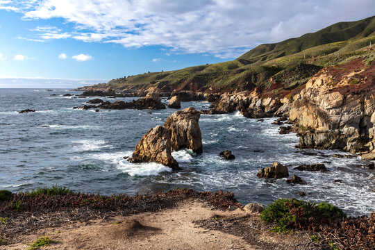 Rocky coastline of Garrapata Beach, south of Monterey, California. Giant rocks just offshore. Waves washing against rocky cliff face. 
