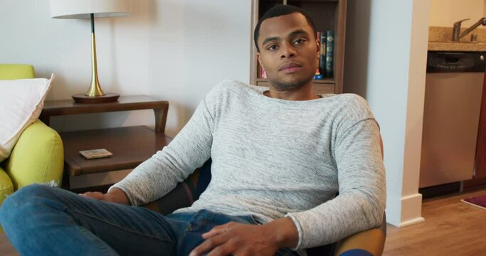 Portrait of attractive young black man sitting on a chair in his apartment. African American Millennial looking at camera wearing jeans and a gray sweater. 4k Slow motion handheld