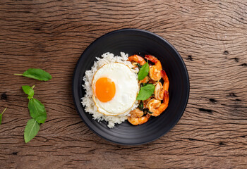 Stir-fried shrimp with basil Thai street food arranged on a black plate with a fried egg Spicy Thai food placed on a wooden table. Top view.
