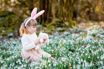 Adorable little girl with Easter bunny ears holding soft plush toy in spring forest on sunny day,...