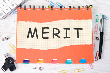 Merit word is written on a sheet in a cage lying on a notebook on the table next to stationery