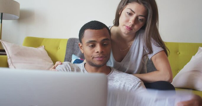 African American and Caucasian couple paying bills online using laptop computer. Millennial newlyweds sitting in living room going over paperwork. 4k Slow motion handheld