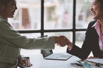 Two Asian businesswomen shake hands to congratulate each other on business success in doing financial business together and investing in the company for future expansion. Startup business idea. - obrazy, fototapety, plakaty