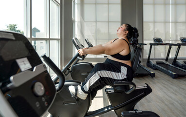 A plus-size lady is energetically riding an exercise bike at a gym, looking focused and determined.