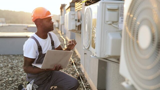 Responsible african american worker repairing device for cooling premises on rooftop with portable laptop in hands. Busy man kneeling near air conditioner during beautiful sunset on background.