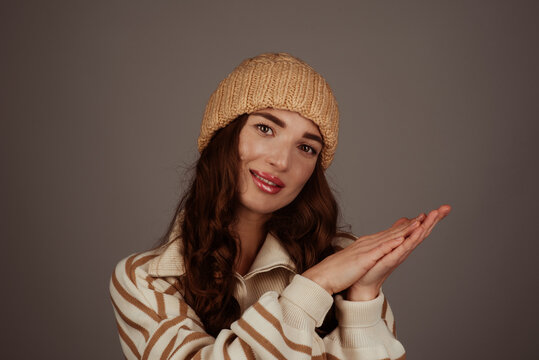 A beautiful girl wearing a beige large-knit hat and sweater on a gray background. Place for text. Winter stylish image