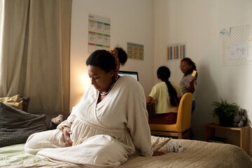 African American pregnant mother sitting on bed and looking at her belly with her daughters in background