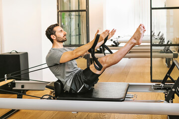 Young man in sportswear doing teaser Pilates exercise and stretching on reformer bed