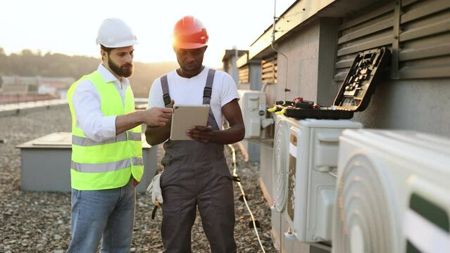 Multiethnic construction managers in protective hard hats reading information from modern tablet about repairing air conditioner. Two men standing on rooftop of industrial factory.