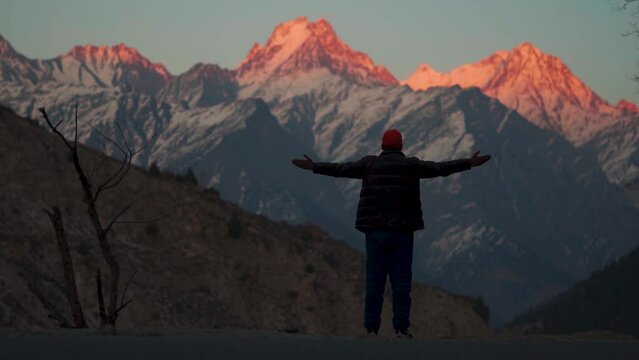 Rear view shot of an Indian man wearing hat and jacket raising arms in front of snow covered Himalayan mountain peaks during the sunset at Kinnaur in Himachal Pradesh, India. Traveler raises his arms