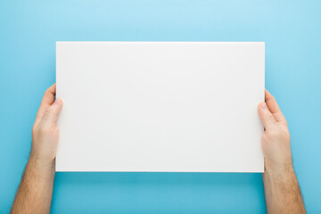 Young adult man hand holding and showing new white ceramic tile on light blue table background. Pastel color. Closeup. Point of view shot. Empty place for text. Top down view. - obrazy, fototapety, plakaty