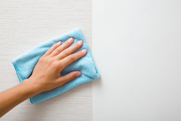 Young adult woman hand holding blue dry rag and wiping surface from glue after light gray wallpaper...