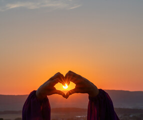 woman creates form a heart shape with hands at sunset. Conveying serene sunset ambiance with heart gesture.