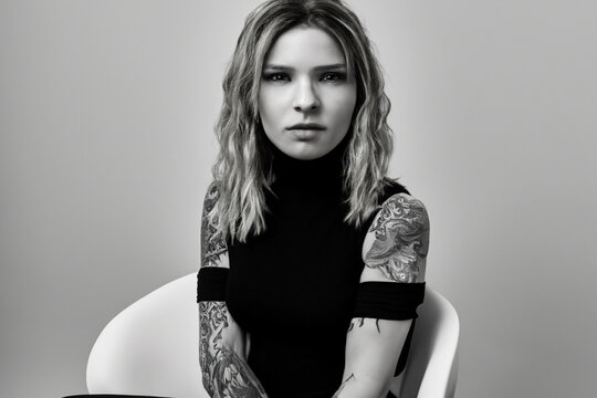 fashionable girl. black and white style photography of a girl sitting on a white chair in a black dress with different tattoos on a gray background