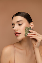 Young pretty female massaging her face by gua sha quartz stone scraper for skincare, posing in studio. Isolated on beige background. Anti-Aging skincare routine