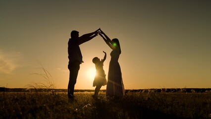 Silhouettes of parents protecting daughter holding hands against sunset on field. Parents hands...