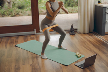 Close up of woman in activewear doing squats with fitness elastic band on mat at home