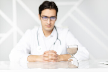 Close-up photo of hourglass on the desktop and male doctor on background. Time and health concept