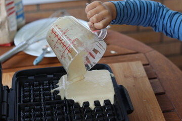 Child making waffle with the waffle machine plate. Child holding cup pouring wet cake mixed batter...