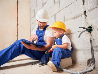 Fototapeta na wymiar Male construction worker sitting by brick wall and pointing at clipboard while showing documents to child. Man and kid wearing safety helmets and work overalls while working on home renovation.