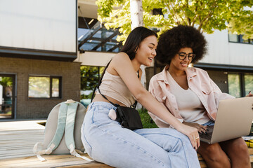 Two positive female students using laptop while studying together outdoors