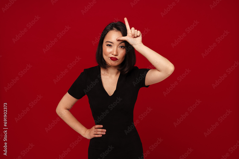 Wall mural Young woman showing loser sign and grimacing isolated over red studio background