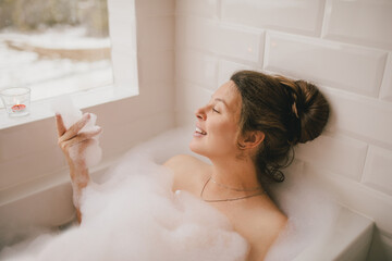 Young woman taking bath with foam near big window.
