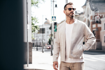Handsome confident stylish hipster lambersexual model. Sexy modern man dressed in elegant white suit jacket. Fashion male posing in the street background in Europe city at sunset. In sunglasses - obrazy, fototapety, plakaty