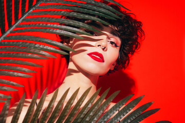 Fashion beauty portrait of young confident brunette woman with evening stylish makeup and perfect clean skin. Sexy model with curly hair posing in studio. With red bright lips. Tropical palm leaf