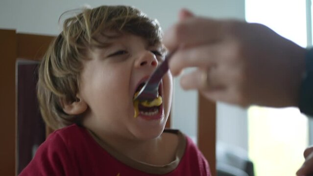 Feeding messy child scrambled eggs during breakfast morning time. Close-up small boy face accident of falling food from mouth