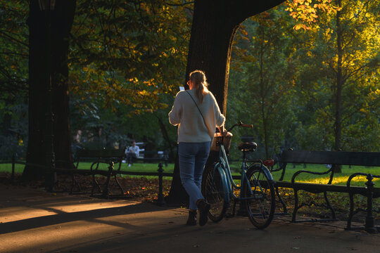 A young woman with a phone walks on a bicycle in a park in the autumn evening, the trees are painted in autumn colors. Peaceful autumn day in the park