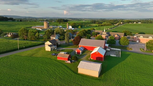 American farm with red barns and green fields. Aerial shot of rural Pennsylvania during summer sunset. Churches and farmland.