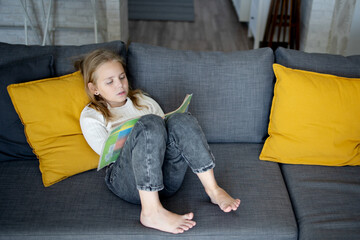 A little blonde girl is intently immersed in the world of adventures, reading an interesting book on the couch at home.