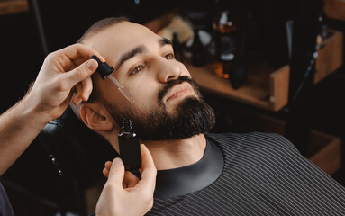 Barber applies beard oil with dropper for man in barbarshop.
