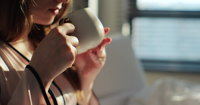 Close up of attractive young woman drinking cup of coffee or tea in the bed and enjoys herself at morning. Calm and cozy morning.