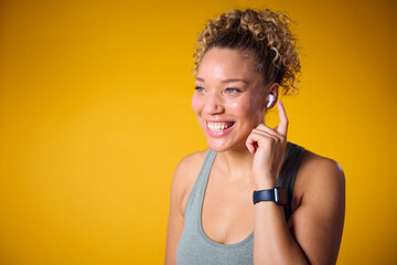 Studio Shot Of Woman In Gym Fitness Clothing Streaming To Wireless Ear Buds On Yellow Background