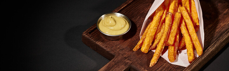 banner of salty crispy French fries inside of paper cone near dipping sauce on wooden cutting board