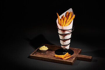 gourmet crispy French fries inside of paper cone near dipping sauce on wooden cutting board