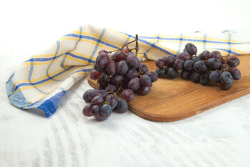 Cutting board with bunch of organic black grapes on white wooden background..