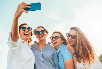 Portrait of four cheerful smiling women in sunglasses embracing together and making selfie photo using modern smartphone during outdoor walk. Woman's friendship, relations, and happiness concept image