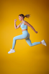 Studio Shot Of Woman Wearing Gym Fitness Clothing In Mid-Air Exercising On Yellow Background