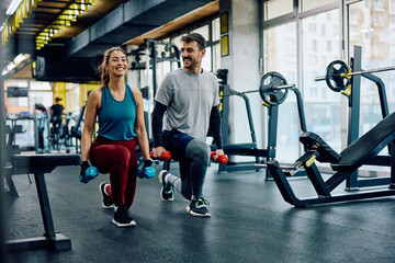 Happy athletic couple exercising with hand weights in lunge position in gym. - obrazy, fototapety, plakaty