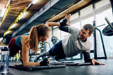 Happy athletic couple giving high five while practicing in plank position in gym.