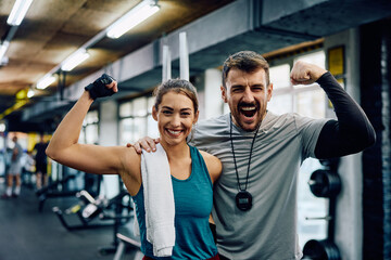 Happy athletic woman and her fitness instructor flexing muscles in gym while looking at camera. - obrazy, fototapety, plakaty