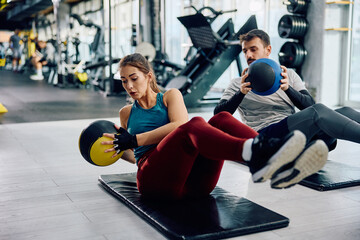 Athletic couple using medicine balls while doing sit-ups during sports training in gym.
