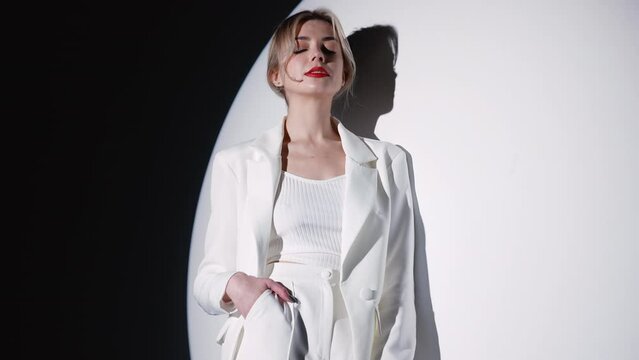 Young beautiful smiling blond woman wearing trendy white suit and jacket. Fashion model posing in studio. Fashionable female posing near white wall in a circle light. Cheerful and happy. With red lips
